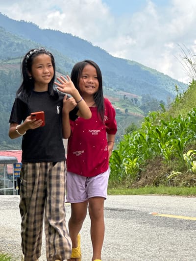 The image captures two young East Asian girls walking on a paved asphalt road in Pung Luong, Vietnam. The girl on the left, with long dark hair and a braided headband, wears a black short-sleeve t-shirt featuring a white polo player logo, plaid wide-leg trousers, and a white beaded bracelet on her left wrist. She holds a red smartphone in her left hand and raises her right hand in a waving gesture, smiling towards the camera. The girl on the right, also with long dark hair, wears a red short-sleeve t-shirt with a repeating white "NIM NIM" pattern, light purple shorts, and yellow quilted sandals. She smiles towards the camera. The background consists of steep, forested mountains under a cloudy sky. To the right, a slope cultivated with rows of young green plants, consistent with corn, is visible. To the left, a metal fence and a partially visible blue sign with white Vietnamese text, including "DON KHAC" and "NOI," are present. The foreground shows the asphalt road with a yellow painted center line and a white reflective road marker.