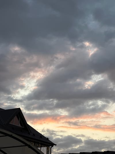 The image captures a cloudy sky with hues of gray, white, and soft pink, suggesting a twilight or overcast day. The lower portion of the frame shows the dark, angled roof of a building, possibly a house or a commercial establishment, with a triangular window visible on the left side. A white awning extends from the building, and string lights are visible beneath it, adding a decorative element. The architecture suggests a residential or possibly a cafe setting. The environment appears to be an outdoor scene, likely in a developed area given the presence of a building with architectural features. There are no people or animals visible in the image. The overall mood is calm and somewhat dramatic due to the cloudy sky. The location is identified as Yaroslavskaya, Russia.