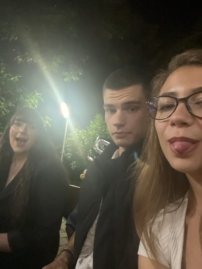 The image is a close-up, dimly lit selfie taken at night, possibly outdoors in Beguntsi, Bulgaria. Three young individuals are visible. On the left, a woman with dark, long hair and glasses is looking at the camera with her mouth open in a surprised or excited expression. In the center, a young man with short dark hair and a neutral expression is looking slightly to the side. On the right, a young woman with long blonde hair and glasses is sticking her tongue out at the camera, her mouth slightly open. She is wearing a white top and a necklace. In the background, there are trees and a bright, vertical outdoor light fixture. Some balloons are also visible in the lower right portion of the frame. The overall impression is of a casual social gathering.