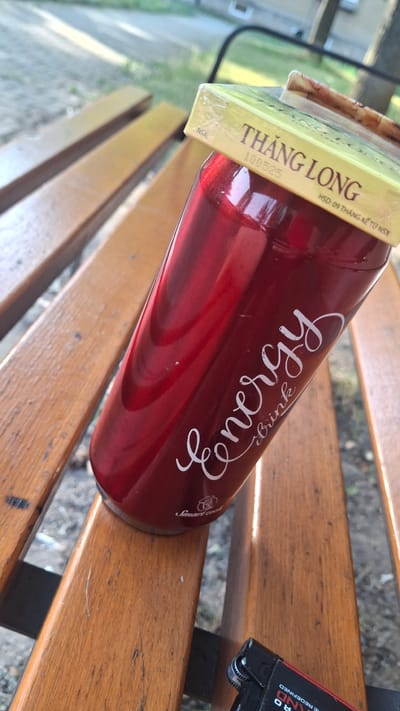 This image shows a close-up of a red energy drink bottle placed on a wooden bench. The bottle has white text on it that reads "Energy" and "Smart cool". A portion of a yellow label is visible on the neck of the bottle, with text including "THĂNG LONG", "100525", and "HSD: 09 THÁNG KỂ TỪ NSX". To the right of the bottle, a black and red lighter is also on the bench, with some text visible on it, including "RO" and "IE REDEFINED". The bench appears to be outdoors, with some greenery and concrete visible in the background. The lighting suggests it might be daytime, possibly with overcast conditions given the diffused light. The overall impression is a casual scene, perhaps someone taking a break outdoors.