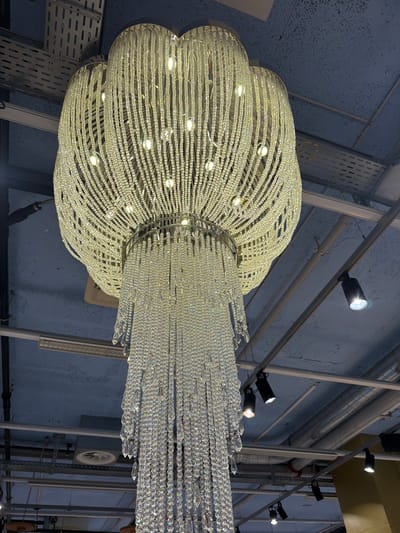 A low-angle, indoor shot shows a large, decorative chandelier hanging from the ceiling. The chandelier consists of numerous strands of beads, arranged in a rounded, flower-like shape at the top and a more rectangular shape below. The strands of beads are illuminated, and light is reflected off of them, creating a sparkling effect. The background reveals a network of metal beams, pipes, and electrical conduits attached to a light blue ceiling. Several track lights are also mounted on the beams, pointed in different directions. The presence of the industrial-style ceiling suggests that the location might be in a commercial or industrial space. There are no people visible in the frame. It is impossible to determine the exact location within Spreitenbach, Switzerland, based solely on the image.