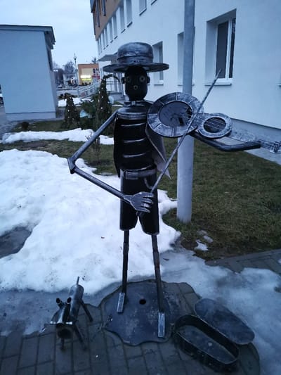 The image displays an outdoor scene in Masty, Belarus, featuring a metal sculpture group positioned on a paved area adjacent to a grassy lawn with melting snow. The central subject is a tall, dark metal sculpture of a stylized human figure, constructed from various metal components. The figure wears a wide-brimmed hat, has bolt-like eyes, and a round mouth opening. Its torso is composed of stacked metal rings, and it wears a rudimentary belt and a cape-like garment. The figure is depicted holding a metal violin-like instrument with its left arm and a metal bow with its right arm, suggesting the act of playing music. The hands are stylized as multi-pronged forks. The figure stands on thin metal legs anchored to a dark, irregularly shaped metal base.
At the base of the main sculpture, to the left, is a smaller, dark metal sculpture of a stylized dog, connected to the main sculpture's base by a metal chain. To the right of the main sculpture's feet, an open, dark metal container, resembling a collection box or instrument case, rests on the pavement.
The ground consists of a dark paved surface under the sculptures, bordered by areas of melting snow and patches of brown and green grass. In the background, a light-colored, multi-story building with multiple windows is visible to the right. A grey pole stands immediately behind the main sculpture. To the left, a smaller, light-colored building with a sloped roof is present. Further in the distance, additional urban structures, streetlights, and sparse vegetation are visible under an overcast, light grey sky.