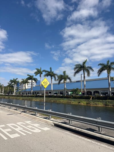 The image displays a sunny, daytime urban landscape in Doral, United States of America, featuring a road, a waterway, palm trees, and buildings under a clear blue sky.

In the foreground, an asphalt road surface is visible, marked with white painted letters that spell "STOP". A grey metal guardrail runs parallel to the road, separating it from a dark, calm waterway or canal. The banks of the waterway are lined with lush green hedges and some taller reeds.

Beyond the waterway, a prominent row of tall, mature palm trees stands between the water and a series of multi-story commercial or residential buildings. These modern buildings feature reflective glass facades and various storefronts. Visible text on building signs includes "MEDICAL CENTER & SPA", "PERFECT VISION CENTER", and a partial "DORAL BANK". Several cars are parked or moving near the buildings.

A bright yellow, diamond-shaped road sign is mounted on a pole near the waterway, clearly indicating "LANE ENDS MERGE LEFT" in black letters. The upper portion of the scene is filled with a bright blue sky, dotted with scattered white cumulus clouds, suggesting clear and pleasant weather. No specific activity or event is taking place; the image captures a static, vibrant scene characteristic of a sunny day in South Florida.