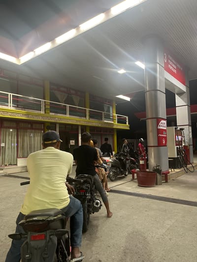 The image depicts a nighttime scene at a Pertamina gas station in Payameuneng, Indonesia. In the foreground, a person wearing a light yellow t-shirt and blue jeans is seated on a motorcycle, facing away from the camera. Another motorcycle is visible behind it, with a person in a black t-shirt also seated on it. Several other individuals and motorcycles are present further back near the gas pumps. The gas station features red and yellow architecture, with fluorescent lighting illuminating the area. A sign on one of the pillars reads "JALUR KHUSUS PERTAMAX" and displays the Pertamina logo. The ground is paved with concrete, and a metal grate runs across a portion of it. The overall impression is one of a busy service station at night.