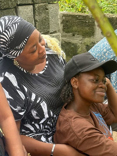 In Ganyingbo, Nigeria, two women are seated outdoors near a brick wall. The woman on the left wears a black and white patterned headscarf and a black top with white patterned accents. She has a leaf-shaped earring. The woman on the right wears a black baseball cap and a brown t-shirt with a graphic design. Both women appear to be looking to their right. The environment includes lush green foliage in the background.
