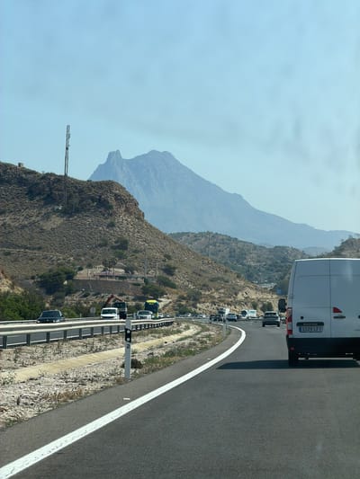 The image depicts a highway scene in Villajoyosa, Spain, under a clear, hazy sky. A white van, possibly a Renault Master based on the visible text on its license plate, is prominently in the foreground, traveling on the right side of the road. The van's license plate reads "3329 LFY". The highway is a multi-lane asphalt road with white lane markings. On the left side of the highway, there is a dry, hilly landscape characterized by sparse vegetation and rocky terrain. A tall communication tower is visible on one of the hills, along with a small structure nearby. In the distance, a large, rugged mountain dominates the horizon, its peak shrouded in a light haze. Several other vehicles, including cars and trucks, are visible on the highway ahead, indicating moderate traffic. The overall impression is a sun-drenched, arid environment typical of the Mediterranean region.