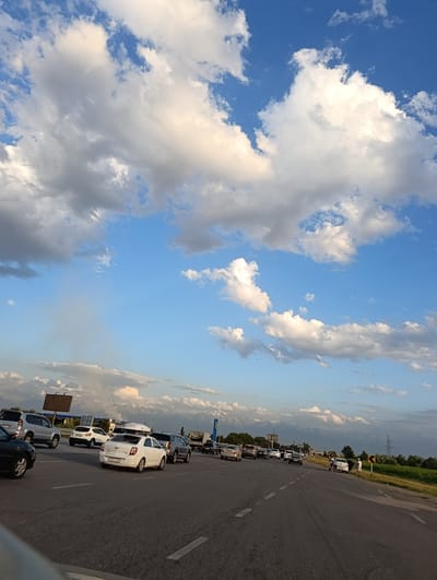 The image captures a traffic jam on a highway with a mountain range in the background. The sky is filled with dramatic, fluffy white and gray clouds against a bright blue backdrop. The road is asphalt with visible lane markings. Several vehicles, including cars and SUVs, are stopped or moving slowly. Some vehicles are white, silver, and dark-colored. There are a few individuals standing on the side of the road near some vehicles. In the distance, trees line the horizon, and a few utility poles are visible. The overall impression is that of a busy highway scene under a partly cloudy sky, potentially on the outskirts of a city like Жаңаталап, Kazakhstan, given the mountainous terrain.