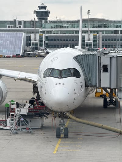 The image shows the nose of a Lufthansa Airbus A350 aircraft connected to a jet bridge at Munich Airport (Flughafen München), located in Oberding, Germany. Airport ground support equipment is present around the aircraft's nose and front landing gear. In the background, the airport terminal building is visible with numbered gates. The scene is overcast, suggesting daylight. No people are visible in the frame.