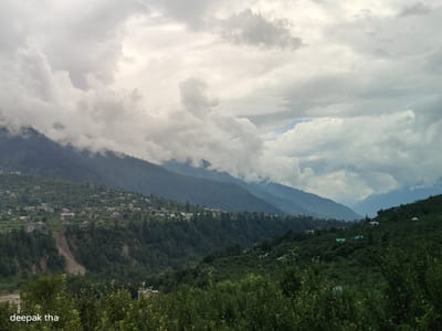 This image depicts a panoramic view of a lush, mountainous landscape with a village nestled on the slopes. The sky is overcast with dramatic, heavy clouds, suggesting a potentially moody or stormy atmosphere. The foreground is dominated by dense green foliage, likely trees and shrubs, which blur slightly, indicating a shallow depth of field or movement.

In the mid-ground, the hillside is dotted with numerous small buildings, characteristic of a village or town. These structures are of various sizes and colors, with some appearing to be traditional houses with pitched roofs, and others possibly more modern constructions. They are interspersed with the greenery of the hillsides, creating a sense of habitation within the natural environment. The slopes themselves are steep and covered in dense vegetation, with some areas showing signs of erosion or landslide scars.

The background consists of layered mountain ranges receding into the distance. The peaks are softened by the atmospheric haze and the overcast sky, with the furthest mountains appearing as muted blue or grey silhouettes.

The text "deepak tha" is superimposed on the lower left corner of the image, likely a watermark or photographer's credit.

While the image shows a village in a mountainous region, it does not provide enough specific visual cues to definitively locate it within the city of Baran, India. The description is a general observation of the scene presented.