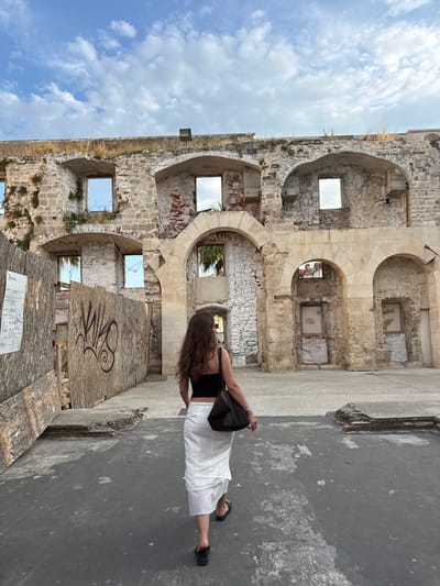 The image shows a person standing on a paved surface in front of ancient stone ruins. The ruins are made of rough-hewn blocks and feature arched openings, some with visible windows. The wall of the structure is weathered, with patches of exposed brick and vegetation growing in some crevices. The ground in the foreground is a dark, textured surface with peeling paint.
The person is viewed from behind, wearing a black top and a long white skirt. They are carrying a black bag over their shoulder and are wearing dark sandals. Their long, dark hair is flowing. The scene appears to be outdoors on a bright, cloudy day, suggested by the clear blue sky with scattered white clouds visible through the openings in the ruins.
The overall impression is one of historical exploration or tourism. The ruins evoke a sense of age and history, possibly part of an ancient palace or city wall. The graffiti on a wooden board leaning against the ruins adds a modern contrast to the ancient stonework. A small informational sign with text and diagrams is visible at the bottom of the frame. The light suggests it is daytime. The location context is Split, Croatia, which aligns with the appearance of ancient Roman or medieval architecture.