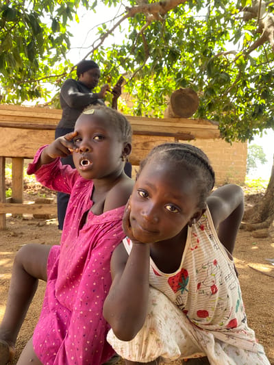 Two young dark-skinned girls are the central subjects of the image. One wears a pink dress with small white hearts, and the other wears a white tank top with strawberry prints. An adult stands in the background with a hijab and is holding a mobile device. The setting appears to be an outdoor area, possibly Hoss, Nigeria, given the location specified. The scene is bathed in sunlight. The environment consists of a makeshift wooden structure, trees with dense foliage, and what appears to be a brick wall. The foreground is sandy and covered in loose dirt.