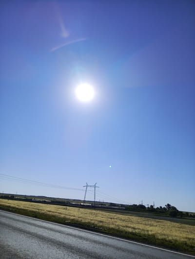 A low-angle, wide shot captures a bright, hazy sun in a clear blue sky, creating lens flare. Below the sky, a vast field of golden-brown grass stretches towards a distant tree line. A high-voltage power line pylon stands in the middle ground, with wires extending across the horizon. In the foreground, a wet asphalt road with visible tire tracks runs diagonally from the lower left to the upper right. The scene appears to be in a rural area, possibly on the outskirts of the city of Studenets, Russia.