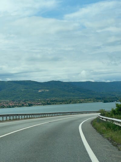 The image captures a panoramic view of a vast body of water, likely a lake or river, bordered by a large mountain range under a partly cloudy sky. The sky is a mix of light blue and white, with diffused clouds. The mountains are covered in dense green vegetation, indicating a lush landscape.
On the right side of the frame, an asphalt road curves gently, following the contours of the shoreline. A solid white line delineates the edge of the road closest to the water. A metal guardrail runs along the side of the road, providing a barrier between the roadway and the water.
Along the base of the mountains, adjacent to the water, several small settlements or villages can be seen, characterized by clusters of buildings with reddish-brown roofs. The scene appears to be set during daytime, with clear visibility and natural light. There are no people visible, and no specific activities are taking place; it is a static landscape shot, possibly taken from a moving vehicle. No text is visible in the image. The location context indicates this is a view from Drobeta-Turnu Severin, Romania, suggesting the body of water is the Danube River.