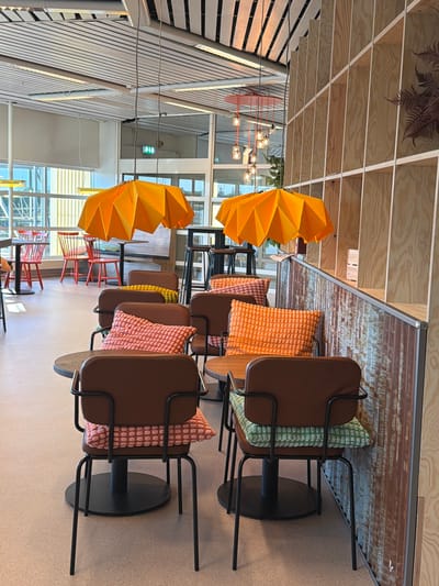This image depicts the interior of a modern cafe, likely located at Malmö-Sturup Airport, Sweden, judging by the presence of a green emergency exit sign. The cafe features a warm and inviting ambiance with natural wood accents and pops of color.
In the foreground, several chairs with brown leather seats and backs are arranged around small, round tables. Each chair is adorned with a decorative cushion: some are pink with a textured pattern, while others are bright orange or light green. The chairs have slender black metal frames and legs, contributing to a contemporary aesthetic.
Dominating the mid-ground are two large, origami-like lampshades in a vibrant orange hue, suspended from the ceiling. These lamps cast a warm glow, complementing the overall cozy atmosphere. Behind the chairs, a wall paneling made of light-colored wood is visible, featuring a grid-like pattern of shelves. A section of this wall has a textured, rustic finish with blue and brown tones, adding visual interest.
In the background, through large windows, glimpses of the outdoors and other parts of the airport terminal can be seen. Red chairs and tables are visible further in the distance, suggesting other seating areas within the cafe or adjacent spaces. The ceiling is modern, with integrated lighting and slatted panels. The floor is a neutral-toned, speckled material, common in public spaces. No people are present in the image, giving the scene a still, observational quality.