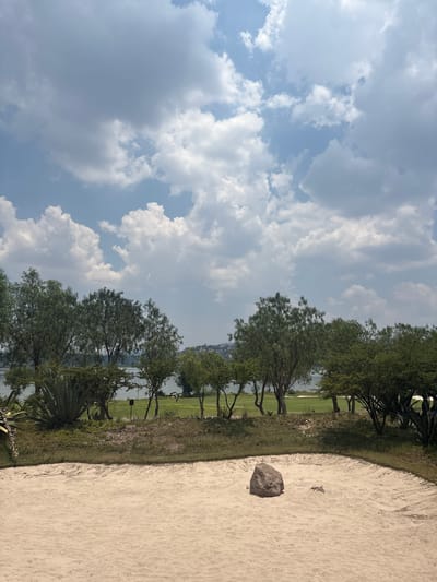 The scene is a sand trap on a golf course in Pueblo Nuevo de Jasso, Mexico. The trap is filled with light-colored sand, and a dark rock is visible within the sand. Beyond the trap, there is a strip of green grass, trees, and a body of water. The sky is cloudy. No people are visible.