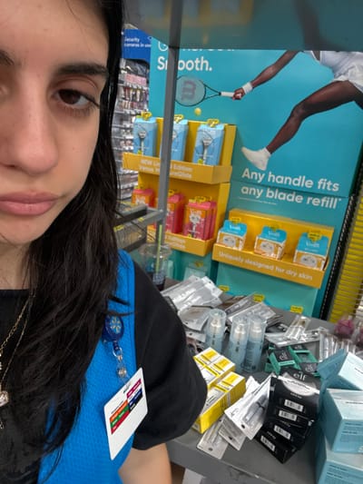 The image captures an indoor scene within a large retail store, specifically a Walmart in Tigard, United States, evidenced by the employee's uniform and the store's typical display elements. The left portion of the frame features a young adult female, visible from the upper nose downwards, with dark hair and a neutral facial expression. She wears a blue uniform vest over a dark long-sleeved shirt. A blue retractable ID badge holder with a yellow starburst logo, consistent with Walmart branding, is clipped to her vest; an illegible ID card with prominent red, orange, green, blue, and purple colored bars hangs from it.
The central and right portions of the image display a cluttered grey counter or table surface laden with various retail products. These include multiple light blue pump bottles, items in clear plastic blister packaging, and numerous boxes of cosmetics or skincare products, notably from the "e.l.f." brand in black and light blue packaging. Several yellow-boxed products are also present. A clear plastic cup containing a dark beverage and a straw rests on the counter amidst the products.
Behind the counter stands a prominent multi-tiered retail display featuring blue and yellow sections. The upper part of the display shows a graphic of a person's lower body and an arm holding a tennis racket with a "WILSON" logo, alongside text that reads "Smooth." and "Any handle fits any blade refill." The lower section of the display includes text stating "Uniquely designed for dry skin." Products visible on the display shelves are "NEW Limited Edition Venus PureGlide" razors or shaving kits in both blue and pink packaging, as well as smaller white boxes labeled "Venus." In the blurred background, additional retail shelving stocked with products is visible, along with a partially obscured sign indicating "Security cameras in use."