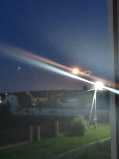 The image shows a nighttime or dusk scene viewed through a window, with a bright light source causing significant glare and lens flare. In the sky, there is a faint rainbow-like arc and a luminous orb, possibly the moon or a bright planet, with some cloud cover around it. Below the sky, a blurry line of buildings with lights on is visible. In the foreground, a street light or similar fixture is emitting a strong white light that beams across the scene. Beyond the light fixture, there is a dark area that appears to be a yard or field with some green grass and indistinct vertical elements that might be fence posts or other structures. The overall image is low resolution and out of focus, making precise identification of objects difficult. The location is identified as Zhal'skoe, Russia.