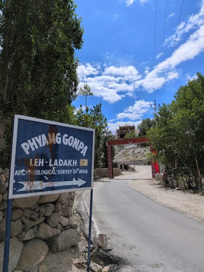 In Phyang, India, a weathered blue sign with white lettering stands to the left of a paved road, indicating "PHYANG GONPA LEH - LADAKH ARCHAEOLOGICAL SURVEY OF INDIA" with an arrow pointing to the right. The sign is mounted on a metal pole and is partially obscured by a low stone wall. The road curves towards a red ceremonial gateway, beyond which a building with golden accents is visible on a hillside. The sky is blue with scattered white clouds, and lush green trees flank the road and the gateway. The scene suggests a peaceful, rural setting on a clear day.