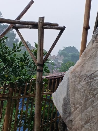 The image presents a serene, verdant landscape, likely set in Welaluhu, East Timor, under an overcast sky. In the foreground, a rustic, weathered bamboo structure dominates the left and center. A prominent vertical bamboo pole anchors a framework of horizontal and diagonal bamboo pieces, creating an open, triangular shape. To the right, another vertical bamboo pole is partially obscured by a large, irregularly shaped object with a rough, light-colored surface, possibly a rock or concrete block, which fills much of the right side of the frame.

Lush, dense green foliage from various trees and bushes occupies the midground, with a bamboo fence running horizontally behind some of the vegetation. In the background, a cluster of residential houses with distinct red-tiled roofs is visible, nestled among rolling, tree-covered hills. A few small, bright white specks, potentially distant lights or reflections, are scattered among these distant structures. The diffused light suggests an overcast day, possibly with mist or light rain, lending a soft, muted quality to the scene. No people or specific activities are visible, emphasizing the tranquil and natural character of the environment. There is no visible text.