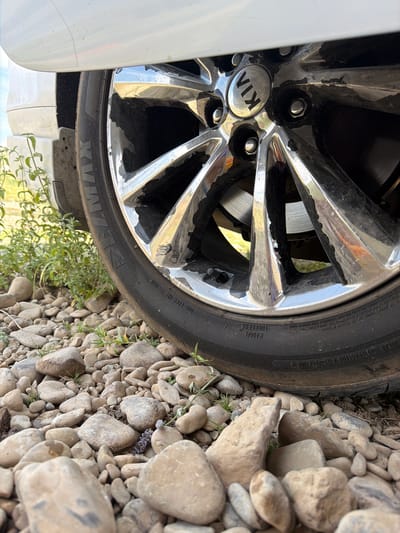 This is a close-up, eye-level outdoor shot of a car tire and rim resting on a rocky surface. The tire is black and partially obscured by the rocky terrain. The rim is silver with a visible "KIA" logo in the center. Some sections of the rim appear to be peeling or damaged. Green plants are visible to the left of the tire. The background shows a blurred landscape, suggesting an outdoor setting. The image does not provide any information about the location in Koktobe, Kazakhstan.