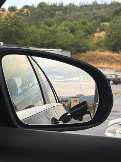 The image is a reflection in a car's side mirror, showing a gas station with a mountain landscape in the background. A black car is parked at the gas pump, and the fuel cap is open. The sky is cloudy, and the sun is setting. The location is a gas station in Levunovo, Bulgaria.