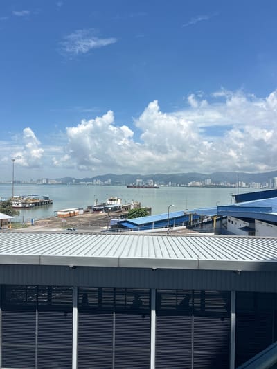 The image shows a wide-angle view of the Butterworth, Malaysia waterfront on a bright, cloudy day. In the foreground, a corrugated metal roof of a modern building extends across the bottom half of the frame, with a dark, louvered facade visible beneath it. Beyond this building, a body of water, likely the Penang Strait, stretches towards the horizon.

Along the waterfront, a port facility is visible with several docked vessels, including passenger ferries and cargo ships. Piers and structures associated with maritime operations are present, with some buildings featuring blue roofs. A city skyline, characterized by numerous buildings and some distant hills, forms the backdrop across the water. The sky is a vibrant blue, punctuated by large, white cumulus clouds. The overall scene depicts a bustling port area with urban development visible in the distance, under a dynamic sky.