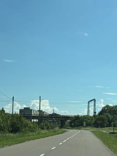 The image captures a paved road curving through a rural or semi-rural landscape under a bright blue sky with scattered clouds. On the left side of the road, a dense green hedge borders the asphalt. In the middle ground, a large, somewhat dilapidated industrial building or complex is visible, supported by a bridge-like structure. Power lines and utility poles are present throughout the scene, indicating infrastructure. To the right of the road, a red rectangular sign with white lettering is visible, along with a diamond-shaped yellow and white road sign. Trees and greenery surround the industrial structure and line the road, suggesting a natural environment juxtaposed with industrial elements. The overall impression is that of a road leading through an area with both natural beauty and remnants of industrial activity. The provided context states the location is in Dobrivliany, Ukraine.