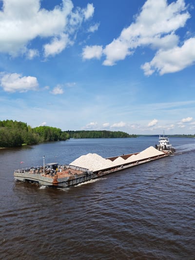 A long barge laden with a large pile of light-colored rocks is being pushed by a tugboat on a wide river. The water is dark brown. Lush green trees line the left bank of the river, while a forested area appears in the distance on the right. A blue sky with scattered white clouds is overhead. A flag is visible on the barge, and there are various pieces of equipment and structures on its deck. This scene may be in Kurikaevo, Russia, although there is no direct, verifiable information to confirm this location.