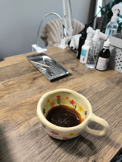 The image depicts an indoor scene in Vinnytski Khutory, Ukraine, likely a professional workstation. In the foreground, a cream-colored ceramic cup, approximately one-third full with a dark brown liquid, rests on a light brown wooden-patterned surface. The cup is adorned with hand-painted floral motifs in red, yellow, and blue. To the left and slightly behind the cup, a rectangular, ribbed silver metal tray contains several small metal tools, consistent with manicure instruments, and a single wooden stick. Further back on the left, a white gooseneck LED desk lamp with a clamp base is visible, and another similar lamp is partially seen behind it. To the right of the tray, a collection of plastic bottles, including spray bottles and dropper bottles with various clear and opaque liquids, are arranged. One amber bottle with a white dropper cap is identifiable with text, partially obscured, appearing to be in Ukrainian. Part of a light grey perforated plastic organizer is visible on the far right. The background features a plain light grey wall, with a light-colored, possibly fur or fleece, item draped over an object behind the lamps. A dark green plant is partially visible in the far right background. No individuals are present in the image, and no active interactions are depicted.