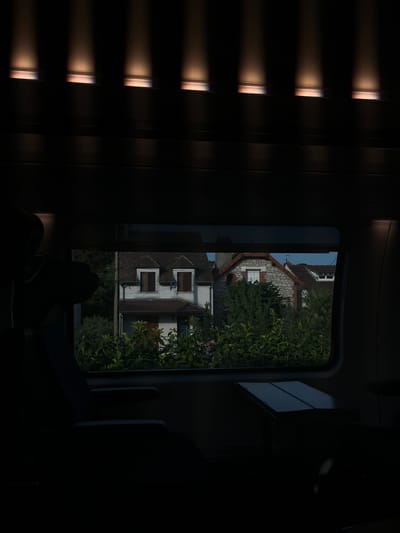 The image captures a view from the window of a moving vehicle, likely a train, looking out at a suburban landscape. The foreground is dominated by the dark interior of the vehicle, with the silhouette of a seat and armrest on the left, and a small fold-out table on the right. The window frames a scene of houses and dense greenery.
Two houses are partially visible. The house on the left appears to be a two-story building with a light-colored facade, brown shutters on its upper windows, and a dark roof. The house on the right is constructed with stone and has a reddish-brown roof with a chimney. Both houses are surrounded by abundant green foliage, with trees and bushes obscuring much of their lower portions. The sky above is a muted blue, suggesting late afternoon or early evening.
The light fixtures visible at the top of the image are integrated into the interior ceiling of the vehicle, casting downward beams of light that are partially diffused by a textured surface. These lights contribute to the overall dark and somewhat moody atmosphere of the interior.
The image does not contain any people or explicit actions. The scene is static, offering a glimpse of a residential area as seen from a passing mode of transport. There is no discernible information within the image that points to the specific location of Saint-Germain-en-Laye, France, beyond what is provided in the prompt.