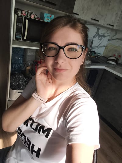 A woman is taking a selfie in her kitchen. She is wearing a white t-shirt with black lettering that reads "TOM HERE". She has on black-rimmed glasses and a pink smartwatch. The kitchen has white cabinets with black handles and a marble-patterned backsplash. In the background, there is a microwave and shelving unit filled with various items. The lighting suggests it is daytime.