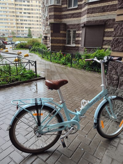 A light blue folding bicycle with a brown leather seat and handlebars is parked on a wet, brick-paved walkway in front of a building with brown and beige brickwork. The bicycle has a rear rack secured with black straps, orange reflectors on the wheels, and a "SHULZ" logo on the frame. To the left of the bicycle, a black metal fence encloses a patch of greenery. In the background, a tall, light-yellow apartment building is visible, along with parked cars and yellow bollards. The scene appears to be in the city of Bugry, Russia, and the wet pavement suggests it has recently rained. There are no people present in the image.