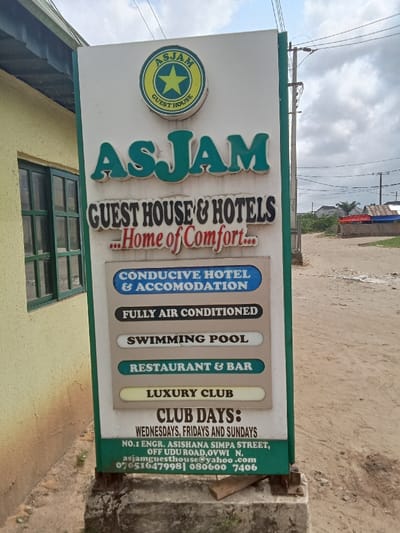 The image shows a sign for a hotel and guest house called "Asjam" located at No.1 Engr. Ashishana Simpa Street, Off Udu Road, Ovwvi, N. The sign lists the services provided: conductive hotel & accomodation, fully air conditioned, swimming pool, restaurant & bar, luxury club, and club days: Wednesdays, Fridays and Sundays. The phone numbers are 07051647998 & 0806007406. The sign is placed on the side of a dirt road. The sign is green, white, and blue.  The sign is on the side of a dirt road in front of what appears to be a brick building with a window and a small wall surrounding it. The background is a rural setting with trees, dirt roads, and a few buildings in the distance. The image is taken from a low angle, and the sign is in focus. The sign is facing the camera and the road is leading away from the camera.