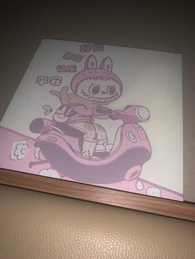 This is a media file depicting a framed print on a wall. The print features a stylized, cartoonish character with large eyes, a wide mouth, and floppy ears, reminiscent of a rabbit. The character is pink and purple, with various numbered sections indicating a paint-by-numbers design. The character appears to be holding a large, abstract object that resembles a hammer or a club.
To the left of the character, there are several lines of text written in what appears to be Chinese characters. The overall aesthetic of the print is playful and whimsical.
The print is framed with a wooden border and is hanging on a textured wall, possibly a leather-textured surface, and a plain grey wall. The lighting suggests an indoor setting, with no indication of time of day or weather. The location context provided is Phú Quốc Special Administrative Region, Vietnam, though there are no specific visual cues in the image to confirm this. The emotion conveyed by the artwork is one of lightheartedness and fantasy.