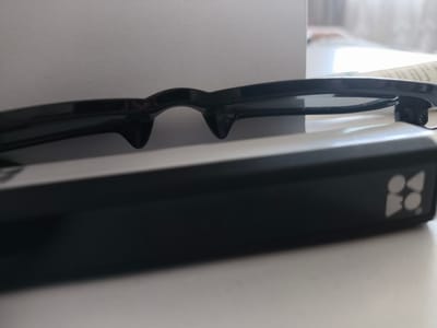 The image depicts a close-up, angled view of a pair of dark-framed glasses resting on a black, rectangular electronic device. The glasses, likely active 3D glasses or smart glasses, feature thick frames with dark, reflective lenses and integrated nose bridge components that extend downwards. They are positioned horizontally, with the front of the frames facing towards the viewer and slightly downwards, obscuring the upper surface of the device beneath them.
The black rectangular device has a smooth, matte finish. On its right end, a light grey or silver logo is visible, composed of stylized geometric shapes: two circles stacked vertically adjacent to two inverted triangles, or alternatively, a cluster of four interconnected circular or rounded elements, preceding what appears to be the letters "BO."
The scene is set indoors. The foreground is a smooth, light grey or off-white surface, likely a table or desk. The background consists of a light grey, subtly textured wall or panel. Towards the upper right of the image, a bright, diffuse light source, possibly a window, casts light onto the scene, creating a soft glow and minimal shadows. No people are visible.