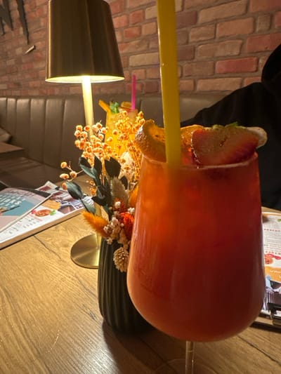 This image depicts an indoor dining or lounge setting in Weißenburg in Bayern, Germany. In the foreground, a stemmed glass contains a red-orange beverage, garnished with a citrus slice and two strawberry slices, and features a yellow plastic straw. To the left of this glass, a black fluted vase holds an arrangement of dried orange, white, and green floral elements. Immediately behind the vase, a golden-toned candle holder with an unlit white candle is visible. A dark brown table lamp with a cylindrical shade casts light from the mid-left. Another beverage in a clear glass with a pink straw and green garnish is partially visible in the mid-background. The surface supporting these items is a light brown wooden table. The background features a textured brick wall above a dark brown upholstered booth seat with horizontal stitching. A person wearing a dark, possibly hooded, garment is partially visible on the far right, seated at the table. A menu or brochure with text and images is partially visible on the table to the far left.