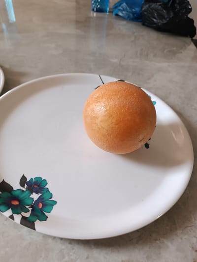 A single, spherical, dull orange object rests centrally on a white, round plate. The object exhibits a finely textured, slightly uneven surface with a distinct sheen, suggesting moisture or an oily coating. Its estimated diameter is approximately one-third the width of the plate. The plate features a decorative floral motif in its lower-left quadrant, consisting of three stylized teal or blue-green flowers with small orange centers, interconnected by dark green/black leaf-like shapes and stems. The plate is positioned on a light gray, tiled, or polished countertop surface, which reflects ambient light and blurred background elements. In the out-of-focus background, a blue plastic object or bag and a black plastic bag are discernible on the same reflective surface. The scene is static, devoid of human presence or activity. The precise location within the city of Anantapur, India, cannot be determined from the visual information provided.