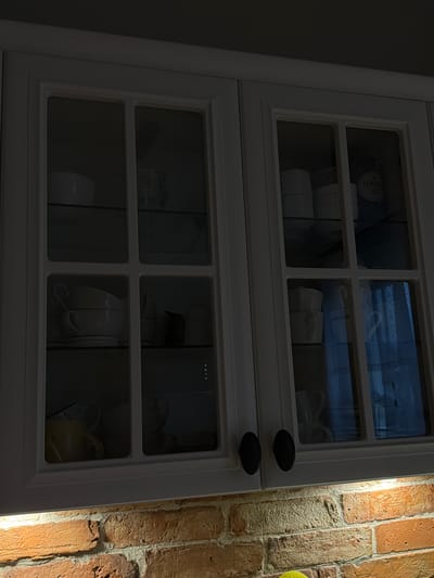The image shows a close-up view of a white cabinet with glass doors, revealing shelves filled with dishes. The cabinet is positioned next to a brick wall, with a warm light illuminating the bricks on the right side of the frame. The dishes appear to be white ceramic, with some cups and bowls visible. In the upper left section of the cabinet, a label with the word "LOVA" is partially visible on a cylindrical object. The overall scene suggests an indoor setting, possibly a kitchen or dining area, with the brick wall providing a rustic or industrial aesthetic. The lighting suggests it is either evening or an indoor setting with artificial lighting. No people are visible in the image.
