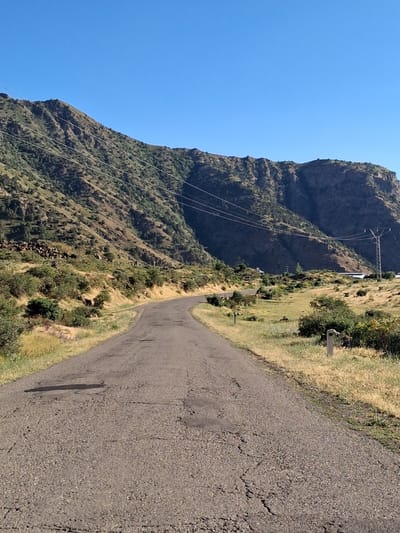 The image depicts a two-lane paved road winding through a mountainous landscape. The road surface is cracked and worn, with a dark stain visible on the left side. The terrain flanking the road is covered in dry, yellow grass and sparse green bushes. In the background, a steep, rocky mountain slope rises, dotted with green vegetation. Power lines stretch across the mountain face, supported by poles. A distant building with a white roof and a transmission tower are also visible on the right side of the frame. The sky is clear and blue. The scene appears to be located in a rural, mountainous area, likely within or near Shatin, Armenia, given the context provided. No people or animals are visible in the image.