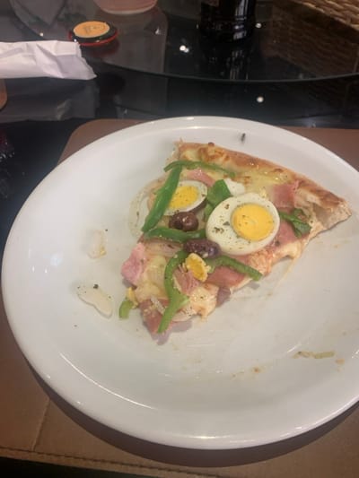The scene depicts an indoor setting, identified as a dining area in Lauro de Freitas, Brazil. A single slice of pizza rests on a white, circular ceramic plate. The pizza slice features a golden-brown crust and a variety of toppings, including melted cheese, two halves of hard-boiled eggs with visible yellow yolks, green bell pepper strips, white onion rings, dark olives (both whole and sliced), and sections of pinkish-red ham. Traces of dried herbs are scattered across the toppings. The white plate exhibits minor food debris and smudges, with two small, white, translucent fragments positioned on its left side. A small, dark-colored insect, consistent with a fly, is present on the upper edge of the pizza crust, near the plate's rim. The plate is situated on a brown surface. The background is out of focus, revealing a dark, reflective surface, likely a glass table, showing light reflections. Partially visible items on the table include a dark cylindrical bottle, a woven basket, a crumpled white paper napkin, and a small, round object with an orange center and black rim. No individuals are depicted, and no specific actions are in progress.