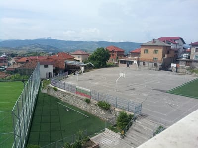 The image depicts an outdoor sports complex with a football field and a basketball court, situated in the town of Valkosel, Bulgaria. The football field, constructed with artificial turf, is enclosed by a tall metal fence. Adjacent to the football field is a basketball court with worn asphalt surfacing and visible court markings. Several basketball hoops are present on the court. A stone wall separates the football field from the basketball court, and a section of the fence above this wall is decorated with the colors of the Bulgarian flag: white, green, and red. Surrounding the sports complex are residential buildings with red-tiled roofs, characteristic of the region. In the background, rolling hills and mountains form the natural landscape. The sky is partly cloudy, suggesting an overcast or changeable weather day. A few people are visible on the basketball court, appearing to be engaged in a casual activity. The overall scene conveys a sense of a community sports facility within a rural or semi-rural setting.