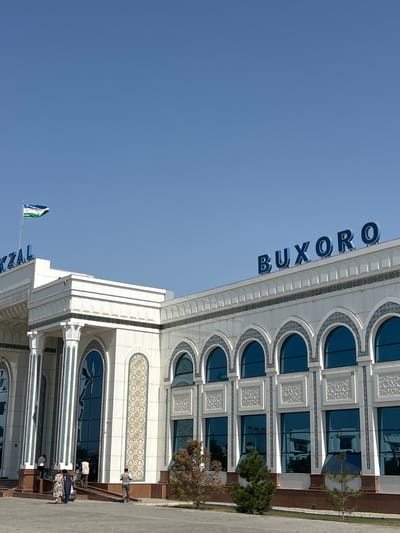 The image depicts the Bukhara-1 railway station in Kagan, Uzbekistan, under a clear, bright blue sky. The prominent building features a multi-story, white marble-like facade adorned with intricate light blue and white traditional Uzbek patterns. At the main entrance on the left, tall fluted columns support an overhang. The right section of the building displays a series of arched windows with dark blue tinted glass, below which are decorative white relief panels. Large blue letters spelling "BUXORO" are affixed to the roofline on the right, while the partial text "KZAL" (likely "VOKZAL") is visible on the roofline to the left. A flag, displaying horizontal stripes of light blue, white, and green with an indistinct emblem on the white stripe, consistent with the national flag of Uzbekistan, flies from a pole atop the left section of the building.
Approximately 8-10 individuals are visible on the paved steps and platform in front of the main entrance. They are dressed in casual attire, engaged in activities such as entering or exiting the building and standing; no distinct interactions are discernible. The foreground consists of a paved area bordering planted sections of soil containing small, scattered evergreen shrubs and trees, some appearing vibrant and others showing signs of dryness. The scene is brightly illuminated by natural daylight, casting defined shadows.