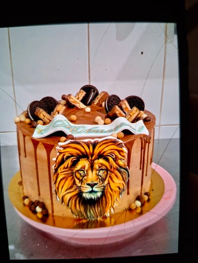 This media file shows a close-up, slightly tilted, top-down view of a decorative cake. The cake is covered in chocolate frosting and adorned with various toppings. Prominently featured on the side of the cake is an image of a lion's head with a golden mane. Around the cake, there are several chocolate sandwich cookies (like Oreos), pieces of candy bars, and small spherical candies in light brown and white colors. The cake rests on a pink and gold cake stand, which is on a white tiled surface. The lighting appears to be artificial, casting soft shadows. There is no discernible text in the image. The scene appears to be indoors, possibly in a home or a bakery, given the tiled surface. The overall presentation suggests it is a celebratory cake, possibly for a birthday or a special event. The image quality is slightly degraded, with visible lines and smudges on the screen capturing the image.