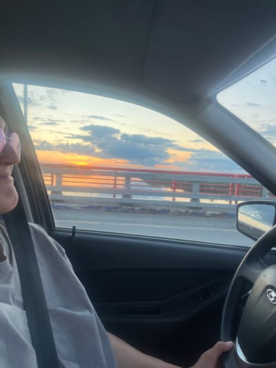 The image shows the interior of a car with a person in the driver's seat, holding the steering wheel. The sun is setting, casting a warm orange glow across the sky, which is partially obscured by clouds. Through the windshield, a bridge or overpass can be seen, with its red railings and white supports. The side mirror reflects the sky and the structure of the bridge. The scene appears to be a peaceful drive during golden hour. The person's arm and shoulder are visible, as is part of their light gray shirt and a black seatbelt. The image is oriented vertically, suggesting it might have been taken in portrait mode. There is no visible text in the image.