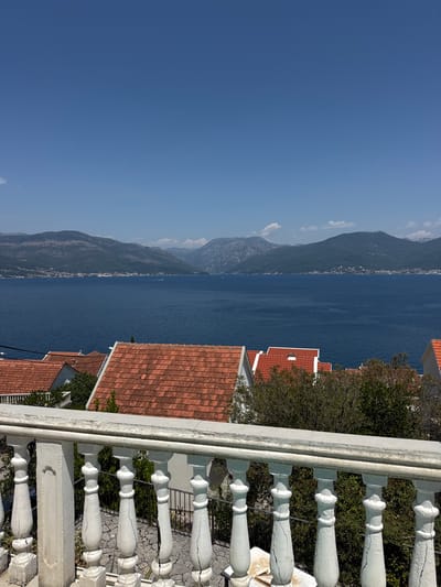 The image shows a scenic view from a balcony or terrace in Krašići, Montenegro. In the foreground, a white, weathered balustrade with decorative balusters is visible, showing signs of age and cracking. Beyond the balustrade, several rooftops with terracotta tiles are spread across a hillside, interspersed with lush green trees and vegetation. In the midground, a wide expanse of deep blue water, likely the Adriatic Sea or a bay, stretches towards a distant mountainous coastline. The mountains are covered in varied shades of green and brown, with some lighter, hazy peaks in the far distance, suggesting a clear, sunny day. A few buildings are visible along the coastline, and small white specks in the water may indicate boats. The sky is a clear, bright blue with a hint of haze towards the horizon. The overall impression is one of a tranquil, sun-drenched coastal landscape.