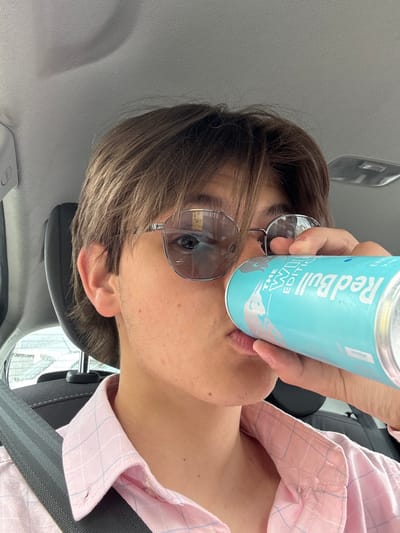 A young man, with dark brown hair and brown eyes, is wearing a pink checkered button-down shirt and a pair of black framed glasses. He is sitting in a car wearing the driver's seatbelt. The man is drinking from a can of Red Bull. He appears to be located in Pocklington, United Kingdom, at a latitude of 53.924 and a longitude of -0.792.