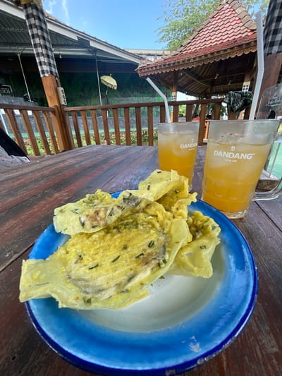 The image depicts a culinary scene likely set in Pandansari, Indonesia, based on contextual clues such as cuisine and surroundings. Two glasses of orange-colored liquid with straws, labeled "Dandang Sejak 1957" are positioned on a wooden table. Adjacent to the drinks is a blue-rimmed plate containing what appears to be fried food, possibly tempeh mendoan, a type of Indonesian fritter. The food has a yellowish hue and green flecks. The backdrop features a partially visible wooden structure, possibly a restaurant or open-air seating area, with a view of the sky and foliage. Additional structures are visible, including roofing and decorative elements, hinting at the local architectural style.
