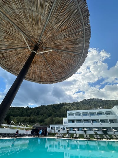 The image shows a view from under a straw beach umbrella towards a building and pool. The sky above is blue with scattered white clouds. The building is white with multiple floors and windows, with green foliage behind. A pool is to the right of the building. Based on the presence of the building, foliage, and pool, this scene is likely located in Faliraki, Greece, a coastal resort town. There is no visible indication of people, actions or interactions.