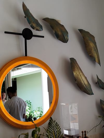 In a well-lit indoor setting, likely a home or hotel in Kendhoo, Maldives, an orange-rimmed circular mirror hangs on a pale wall adorned with decorative leaf-shaped art pieces. The mirror reflects a man wearing a white shirt and glasses, seen from behind, with a woman beside him. Beyond them, lush green plants are visible, suggesting an outdoor area with natural light. A clear acrylic container sits on a surface below the wall art, and further to the right, some wooden objects and a plant with striped leaves are present. The overall ambiance appears relaxed and peaceful. The time of day seems to be daytime, judging by the bright natural light illuminating the scene. There is no visible text in the image.
