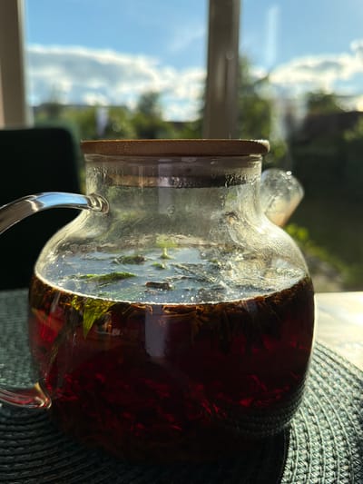 A close-up, eye-level shot captures a glass teapot filled with dark red tea and green mint leaves, its bamboo lid slightly ajar. The teapot sits on a dark green woven placemat on a table, with sunlight streaming in from a window behind it, illuminating the condensation on the glass and the steam rising from the tea. The background is blurred, showing a vibrant blue sky with white clouds, green trees, and hints of buildings, suggesting an outdoor or garden view from a residence in Kerro Massiv, Russia. The overall lighting is bright and natural, emphasizing the freshness of the tea and the pleasant weather.